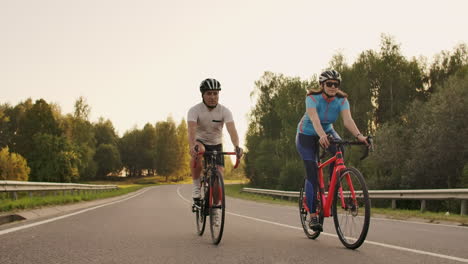 Ein-Mann-Und-Eine-Frau-Fahren-Bei-Sonnenuntergang-In-Zeitlupe-Gemeinsam-Auf-Fahrrädern-Die-Straße-Entlang.-Das-Paar-Ist-Mit-Dem-Fahrrad-Unterwegs-Und-Trägt-Fahrradhelme.